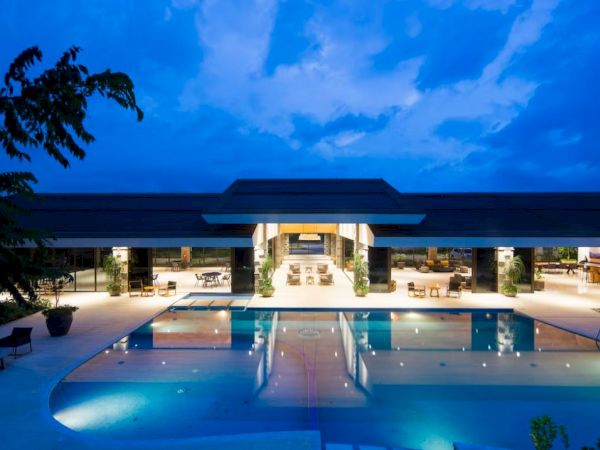 The image shows a luxurious modern building with a lit-up interior, adjacent to a large swimming pool, under a twilight sky.