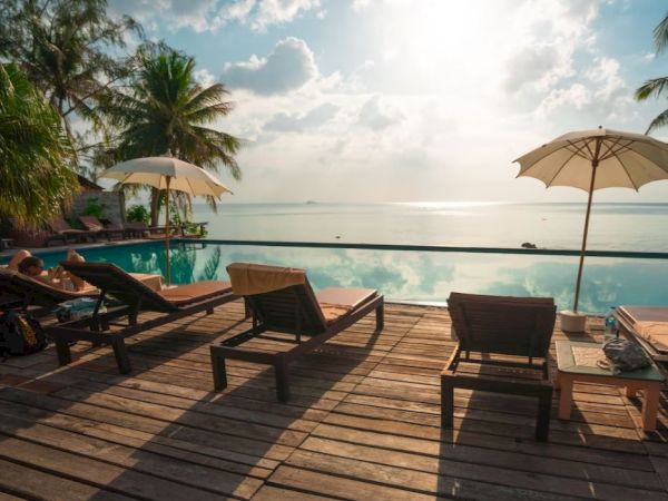 A serene poolside scene with lounge chairs, umbrellas, palm trees, and a view of the ocean under a partly cloudy sky.