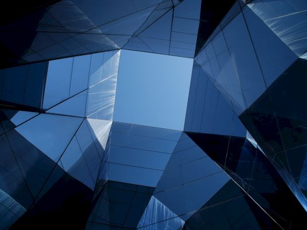 A view looking up through a geometric structure of mirrored glass, reflecting a deep blue sky in the center.