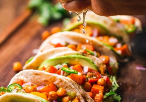 Soft tacos filled with chickpeas, diced vegetables, and avocado slices are being topped with fresh lime juice, on a wooden board.