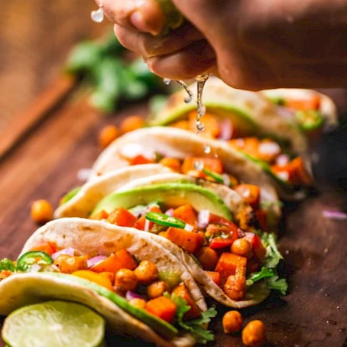 Soft tacos filled with chickpeas, diced vegetables, and avocado slices are being topped with fresh lime juice, on a wooden board.