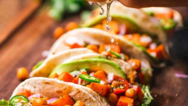 Soft tacos filled with chickpeas, diced vegetables, and avocado slices are being topped with fresh lime juice, on a wooden board.