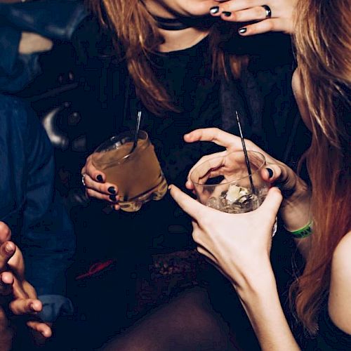 People are enjoying drinks at a social gathering. Laughter and conversation are evident, with cocktails in hand and a lively atmosphere.