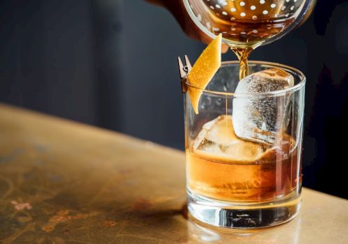 A drink, possibly a cocktail, is being poured into a glass with ice and a lemon peel garnish on a bar surface.