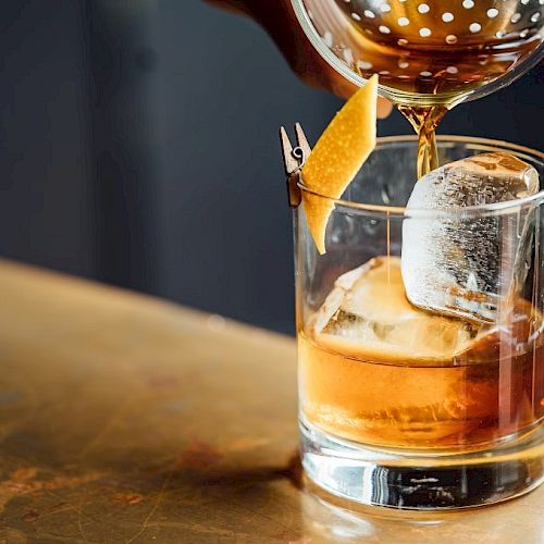 A drink, possibly a cocktail, is being poured into a glass with ice and a lemon peel garnish on a bar surface.