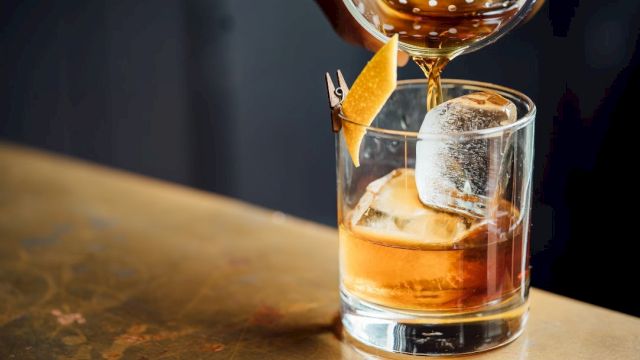 A drink, possibly a cocktail, is being poured into a glass with ice and a lemon peel garnish on a bar surface.
