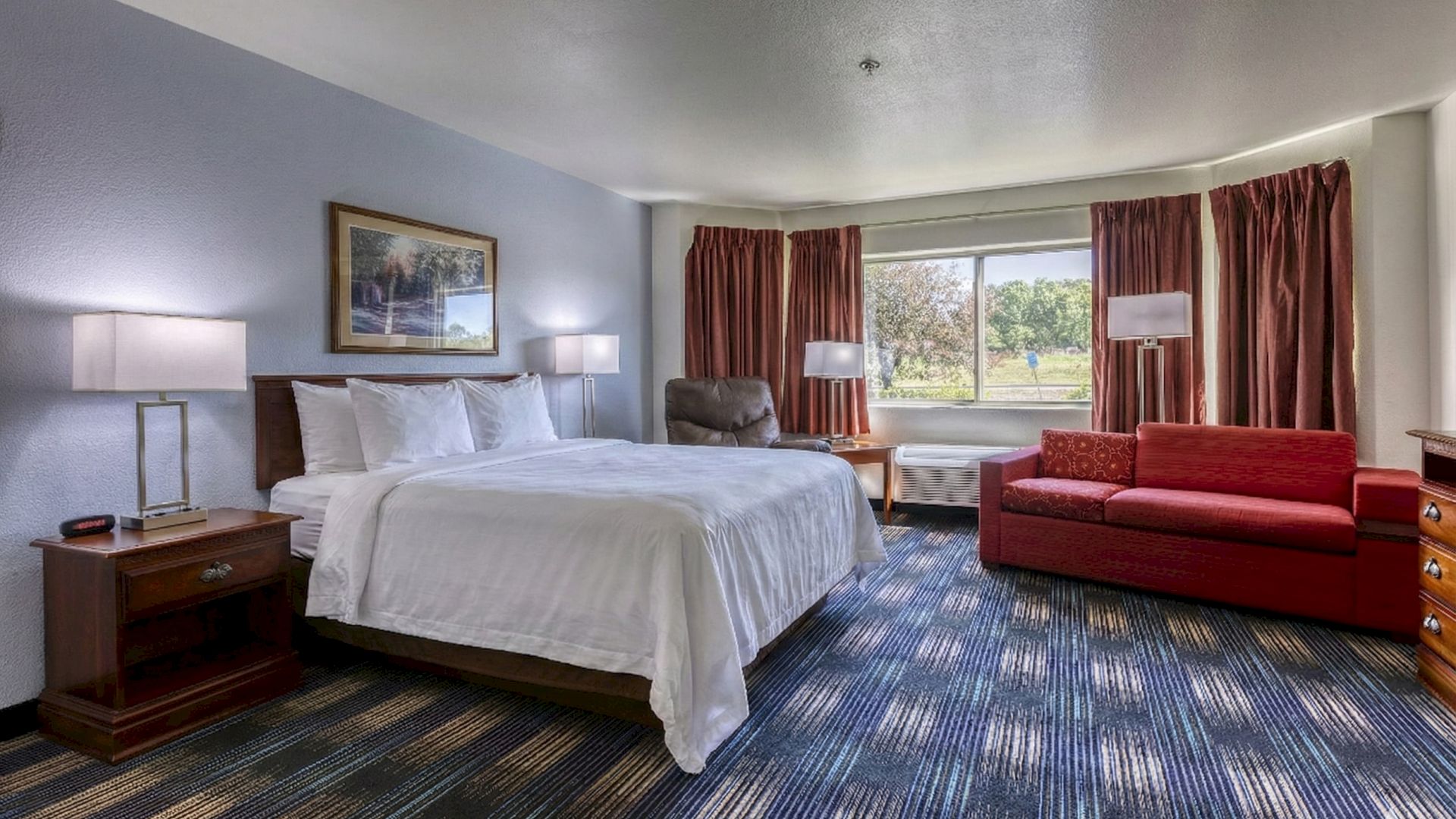 A hotel room with a large bed, nightstands, lamps, a red sofa, two chairs, a window with curtains, and blue patterned carpet, cozy and spacious.