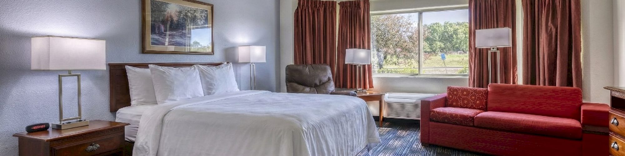 A hotel room with a large bed, nightstands, lamps, a red sofa, two chairs, a window with curtains, and blue patterned carpet, cozy and spacious.