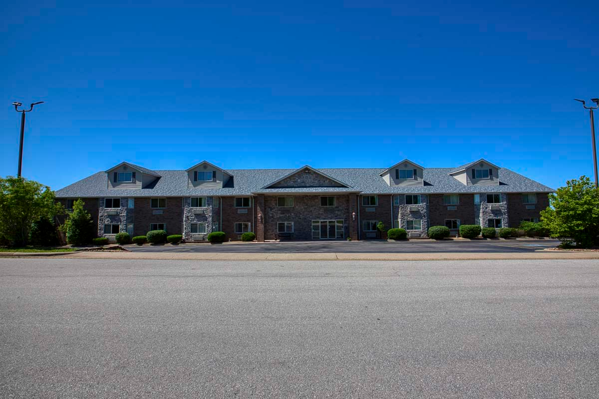 A two-story brick building with a pitched roof and dormer windows, set against a clear blue sky, seen from across a wide pavement.