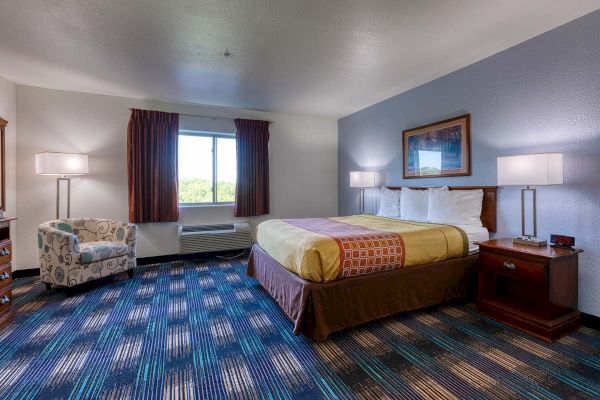 The image shows a hotel room with a double bed, colorful bedding, two side lamps, a chair, a window, and a digital alarm clock.
