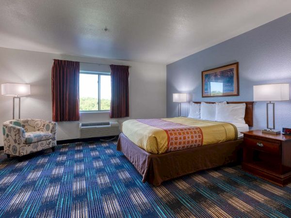 The image shows a hotel room with a double bed, colorful bedding, two side lamps, a chair, a window, and a digital alarm clock.
