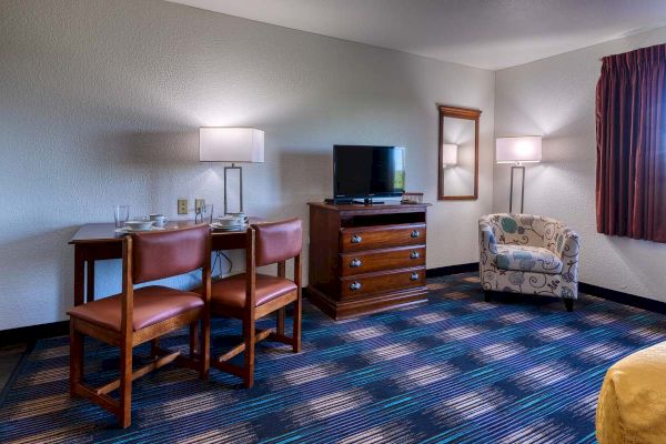 A hotel room with a TV on a dresser, a table with chairs, a decorative armchair, and two floor lamps.
