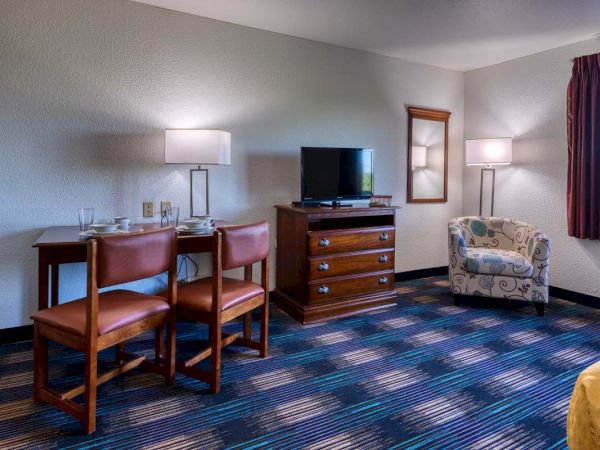 A hotel room with a TV on a dresser, a table with chairs, a decorative armchair, and two floor lamps.