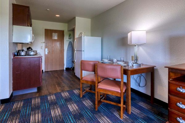 A small hotel room kitchenette with a fridge, stove, microwave, dining table set with dishes, chairs, a lamp, ironing board, and wooden cabinetry.