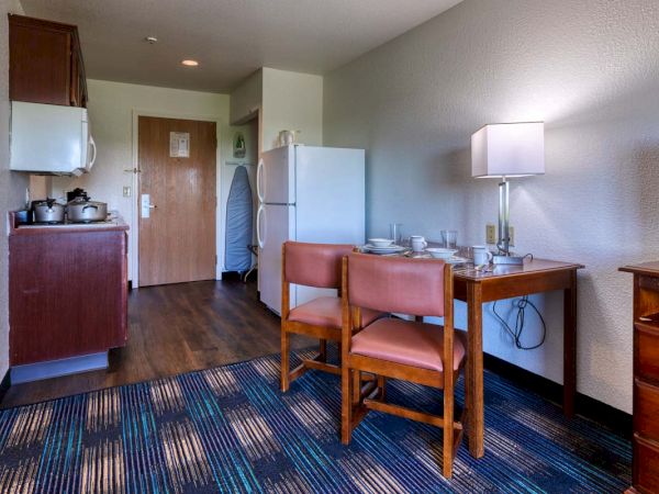 A small hotel room kitchenette with a fridge, stove, microwave, dining table set with dishes, chairs, a lamp, ironing board, and wooden cabinetry.