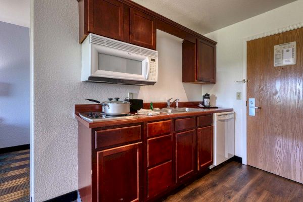 A compact kitchenette with wooden cabinets, a microwave, stove, pot, sink, dishwasher, and a door with a security latch.