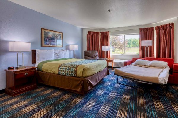 The image shows a hotel room with a bed, a sofa bed, lamps, artwork on the wall, and a large window with curtains.
