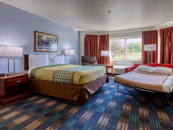 The image shows a hotel room with a bed, a sofa bed, lamps, artwork on the wall, and a large window with curtains.