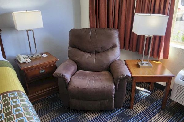 A cozy room features a recliner, two square lamps, bedside tables, a bed, phone, and curtains with a view of greenery outside.