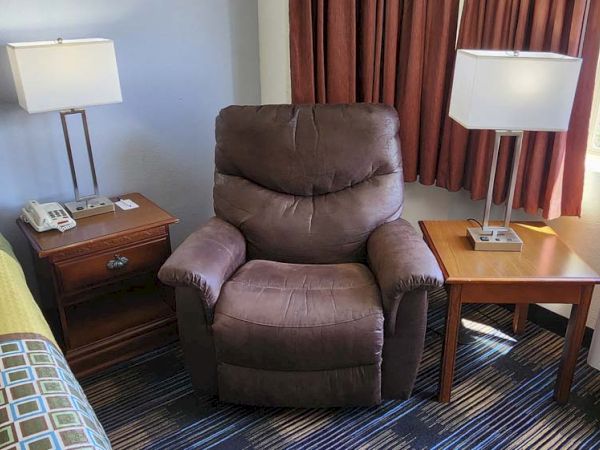 A cozy room features a recliner, two square lamps, bedside tables, a bed, phone, and curtains with a view of greenery outside.