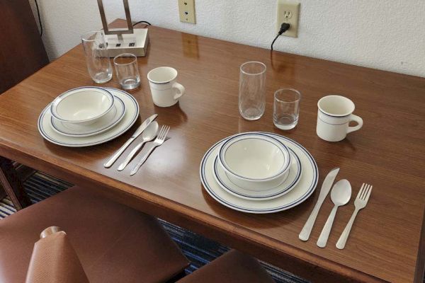 A wooden table set for two with plates, bowls, utensils, cups, and glasses beside a lamp and power outlet on the wall.