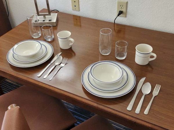 A wooden table set for two with plates, bowls, utensils, cups, and glasses beside a lamp and power outlet on the wall.