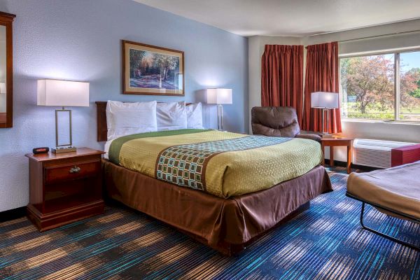 The image shows a hotel room with a bed, armchair, side tables, lamps, a window, and framed artwork on the wall.