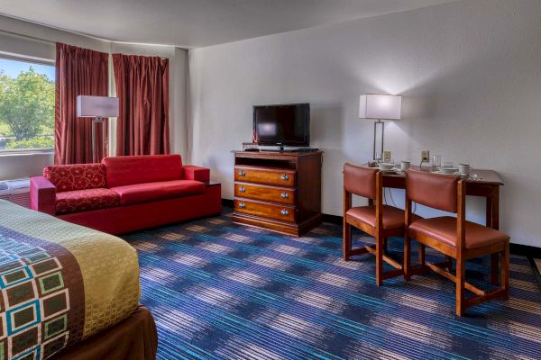 The image shows a hotel room with a red sofa, TV on a dresser, and two chairs at a table with lamps, beside a large window.