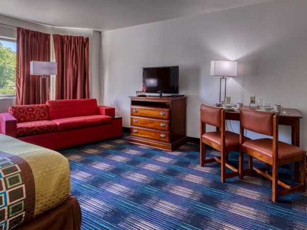 The image shows a hotel room with a red sofa, TV on a dresser, and two chairs at a table with lamps, beside a large window.