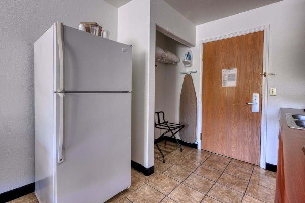 The image shows a kitchenette area with a refrigerator, ironing board, iron, luggage rack, and a closed wooden door.