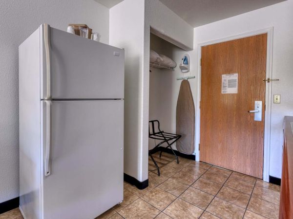 The image shows a kitchenette area with a refrigerator, ironing board, iron, luggage rack, and a closed wooden door.