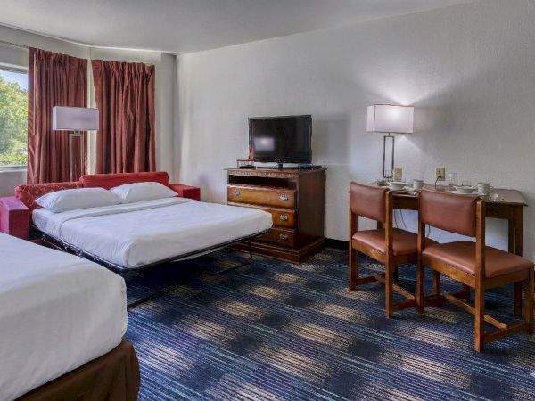 A hotel room with two beds, a red sofa, a dresser with a TV, a desk with two chairs, and a window with red curtains.