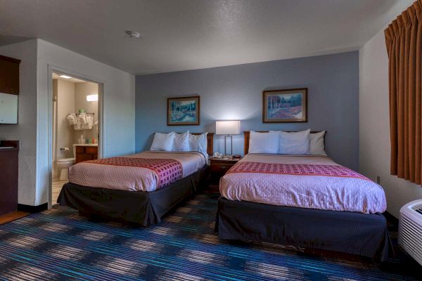 The image shows a hotel room with two beds, a nightstand with a lamp, paintings, and a view of a bathroom.