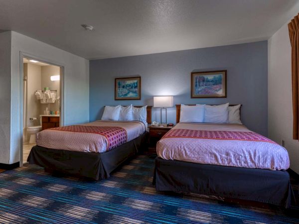 The image shows a hotel room with two beds, a nightstand with a lamp, paintings, and a view of a bathroom.
