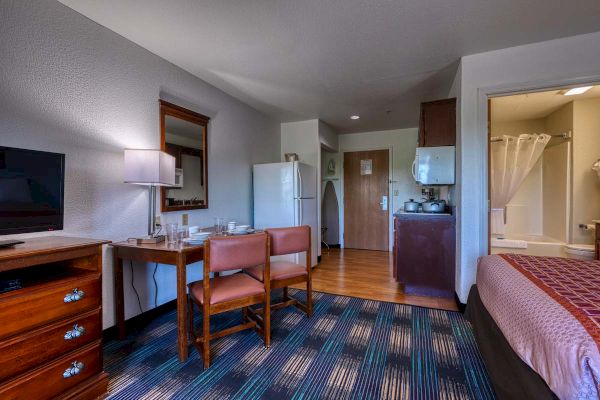 A cozy hotel room with a table, chairs, fridge, TV, and small kitchenette. A bed and bathroom are visible in the corner.