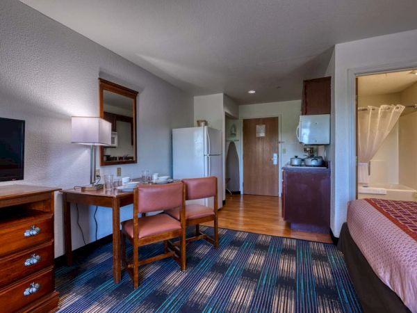 A cozy hotel room with a table, chairs, fridge, TV, and small kitchenette. A bed and bathroom are visible in the corner.