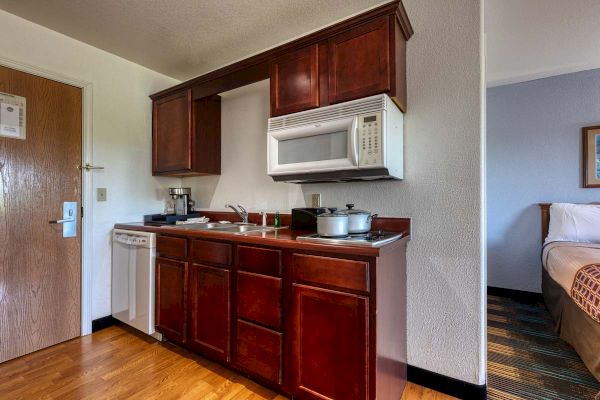 The image shows a small kitchenette with wooden cabinets, a microwave, sink, coffee maker, electric stove, and cooking pots.