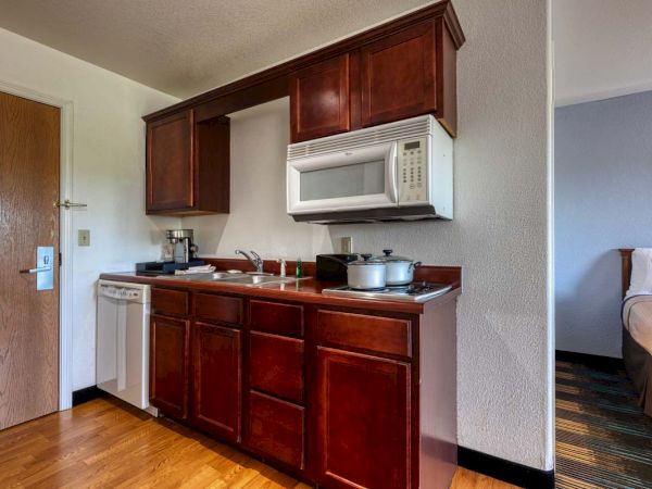 The image shows a small kitchenette with wooden cabinets, a microwave, sink, coffee maker, electric stove, and cooking pots.