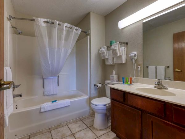 A clean bathroom with a shower, sink, toilet, towels, hairdryer, toiletries, and a large mirror under bright lighting.
