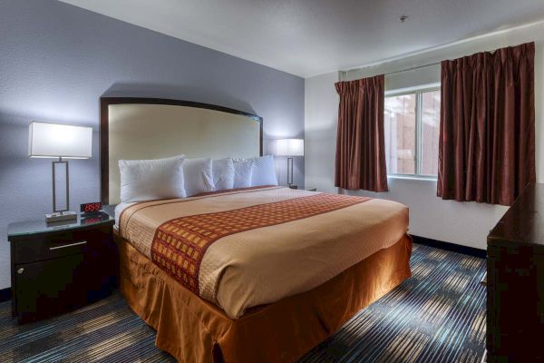 A hotel room with a king-size bed, two bedside lamps, an alarm clock, and a window with curtains.