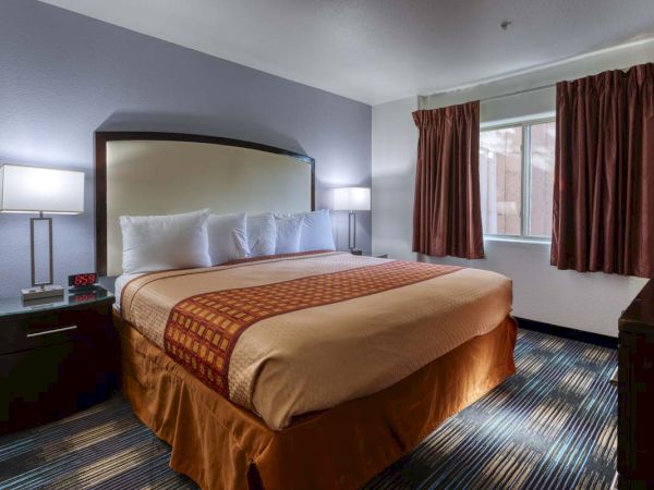 A hotel room with a king-size bed, two bedside lamps, an alarm clock, and a window with curtains.