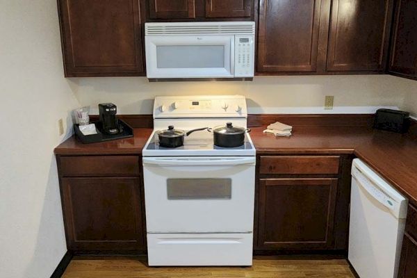 The image shows a kitchen with wooden cabinets, a stove with pots, a microwave, a coffee maker, and a dishwasher.