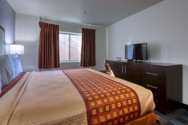 A neatly made hotel room with a patterned bedspread, TV on a dresser, and curtains partially drawn over the window.
