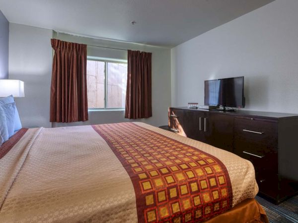 A neatly made hotel room with a patterned bedspread, TV on a dresser, and curtains partially drawn over the window.
