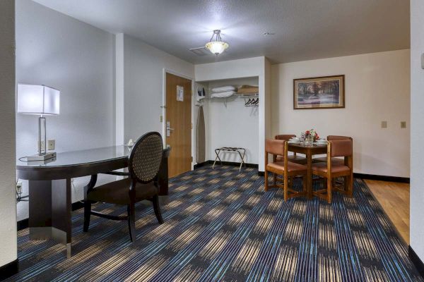 A hotel room corner with a desk, chair, lamp, small dining table with chairs, closet area, ironing board, and a framed painting on the wall.