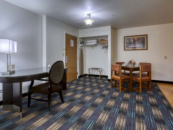A hotel room corner with a desk, chair, lamp, small dining table with chairs, closet area, ironing board, and a framed painting on the wall.