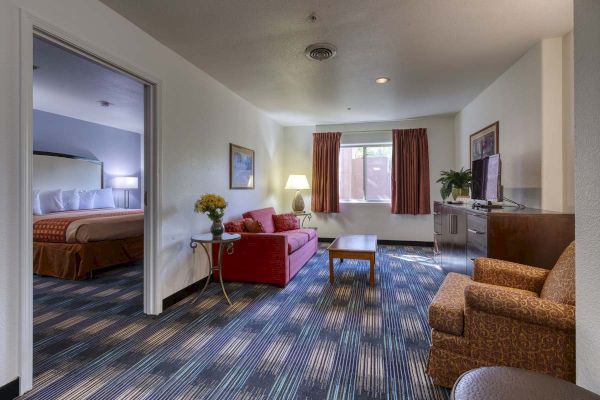 This image shows a hotel room with a seating area, a red sofa, a TV, and a partially visible bedroom through a doorway with a bed.