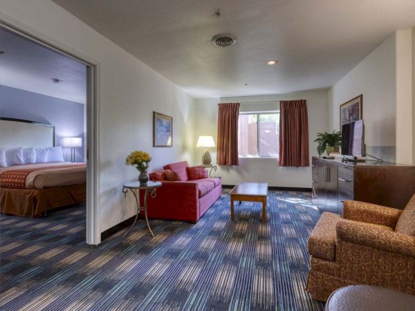 This image shows a hotel room with a seating area, a red sofa, a TV, and a partially visible bedroom through a doorway with a bed.