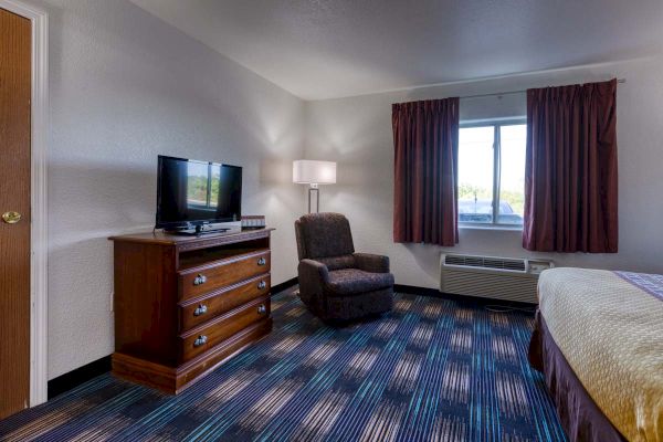 A hotel room with a TV on a wooden dresser, a chair, a lamp, and a window with curtains. There’s a bed partially visible on the right.