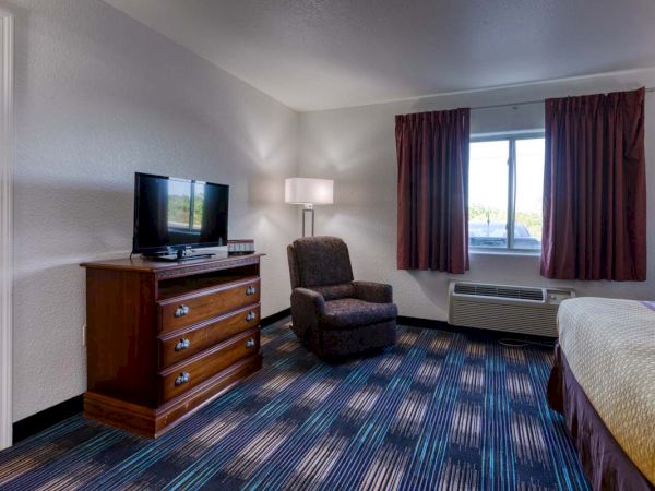 A hotel room with a TV on a wooden dresser, a chair, a lamp, and a window with curtains. There’s a bed partially visible on the right.